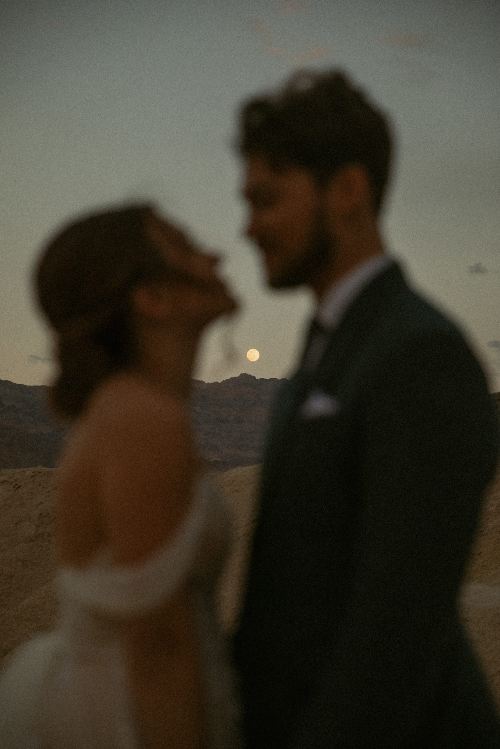 A couple on their elopement day in Death Valley National Park in California, out of focus but looking at each other, and the full bright moon behind them is in focus