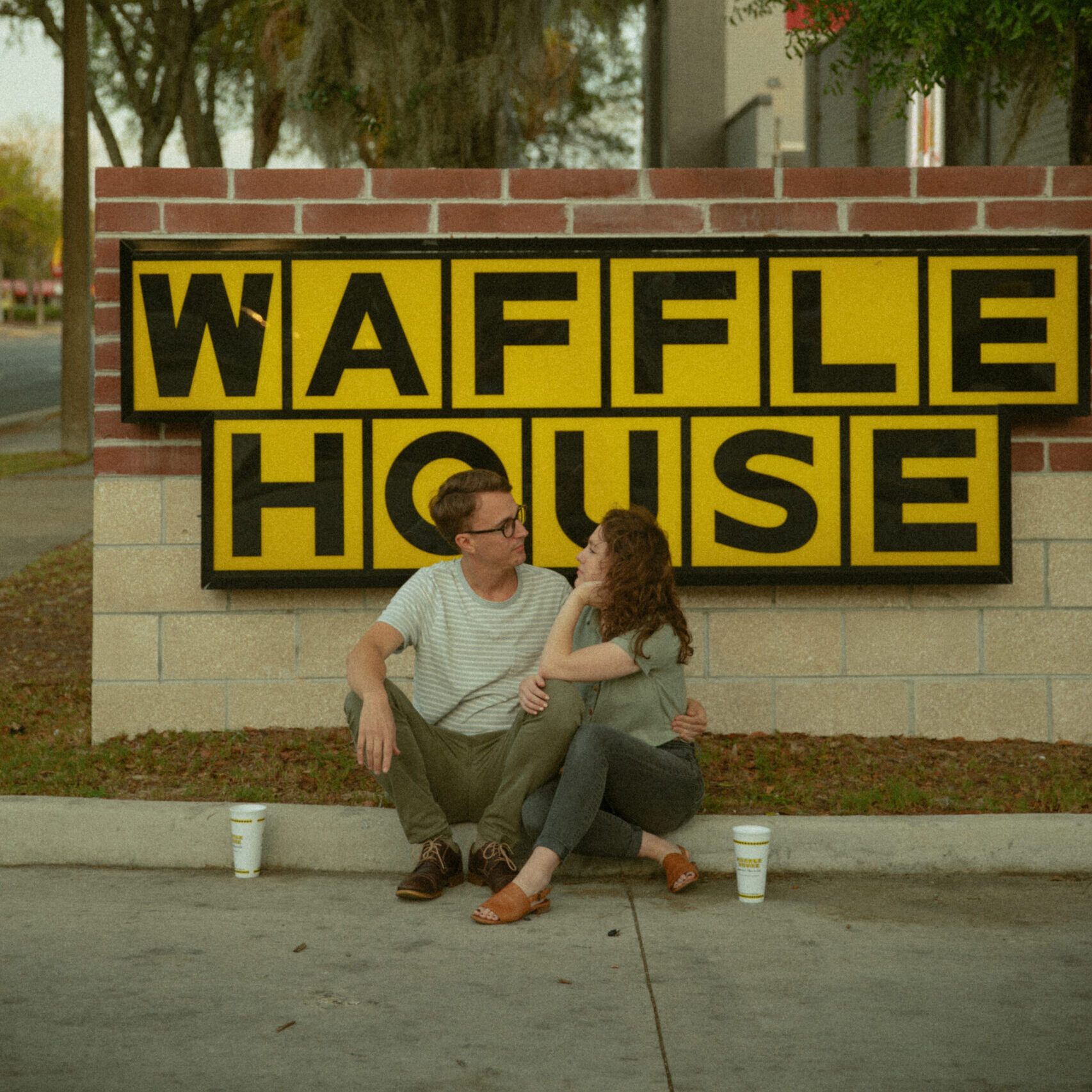 Couple sitting on a curb, looking toward each other, in front of a yellow block letter waffle house logo sign in the parking lot of a restaurant in Gainesville, Florida